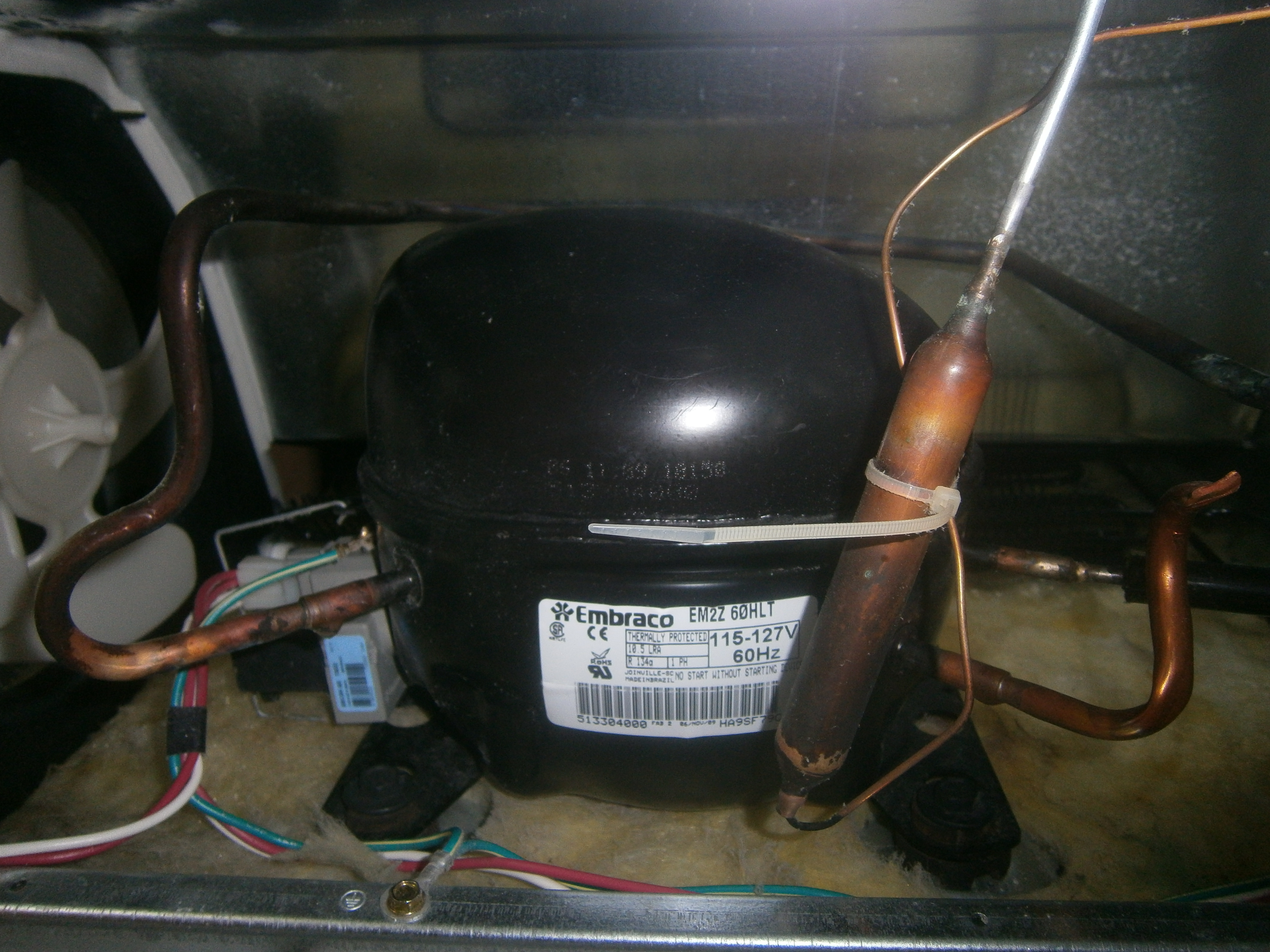 Refrigerator compressor installed beside a fan, with connected wiring and copper refrigerant tubing.