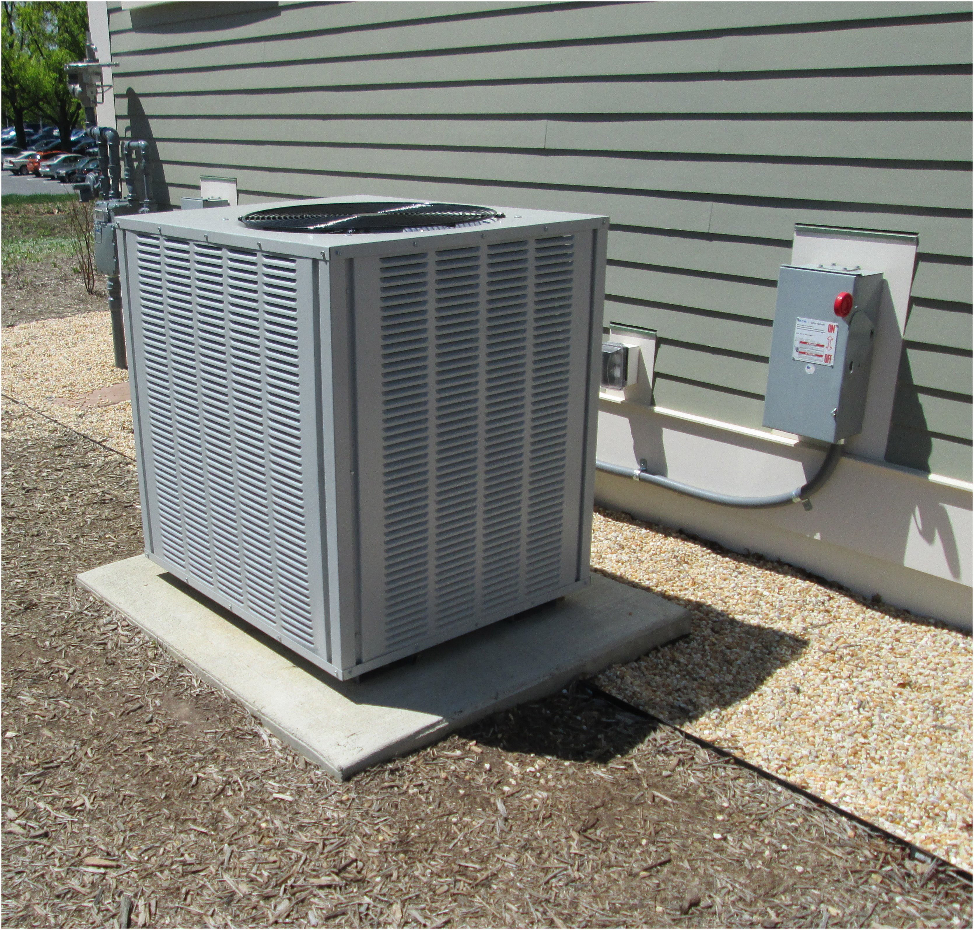 Outdoor air-conditioning unit installed beside a building, with a wall-mounted electrical disconnect and connecting conduit.