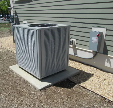 Outdoor air-conditioning unit installed beside a building, with a wall-mounted electrical disconnect and connecting conduit.
